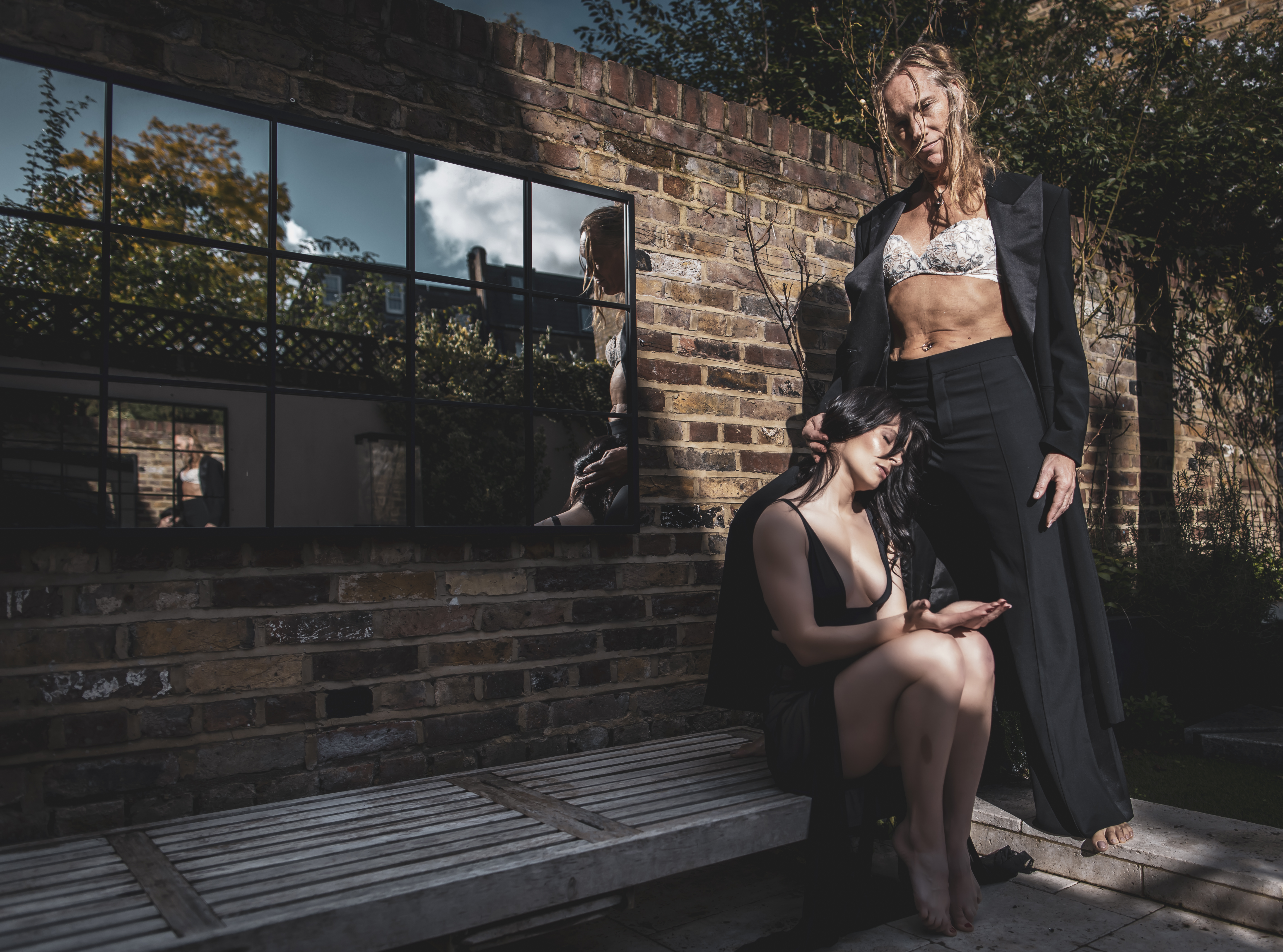 A dominant woman stands calmly over a kneeling submissive, her hand resting on their head — an image of quiet power and contained desire.