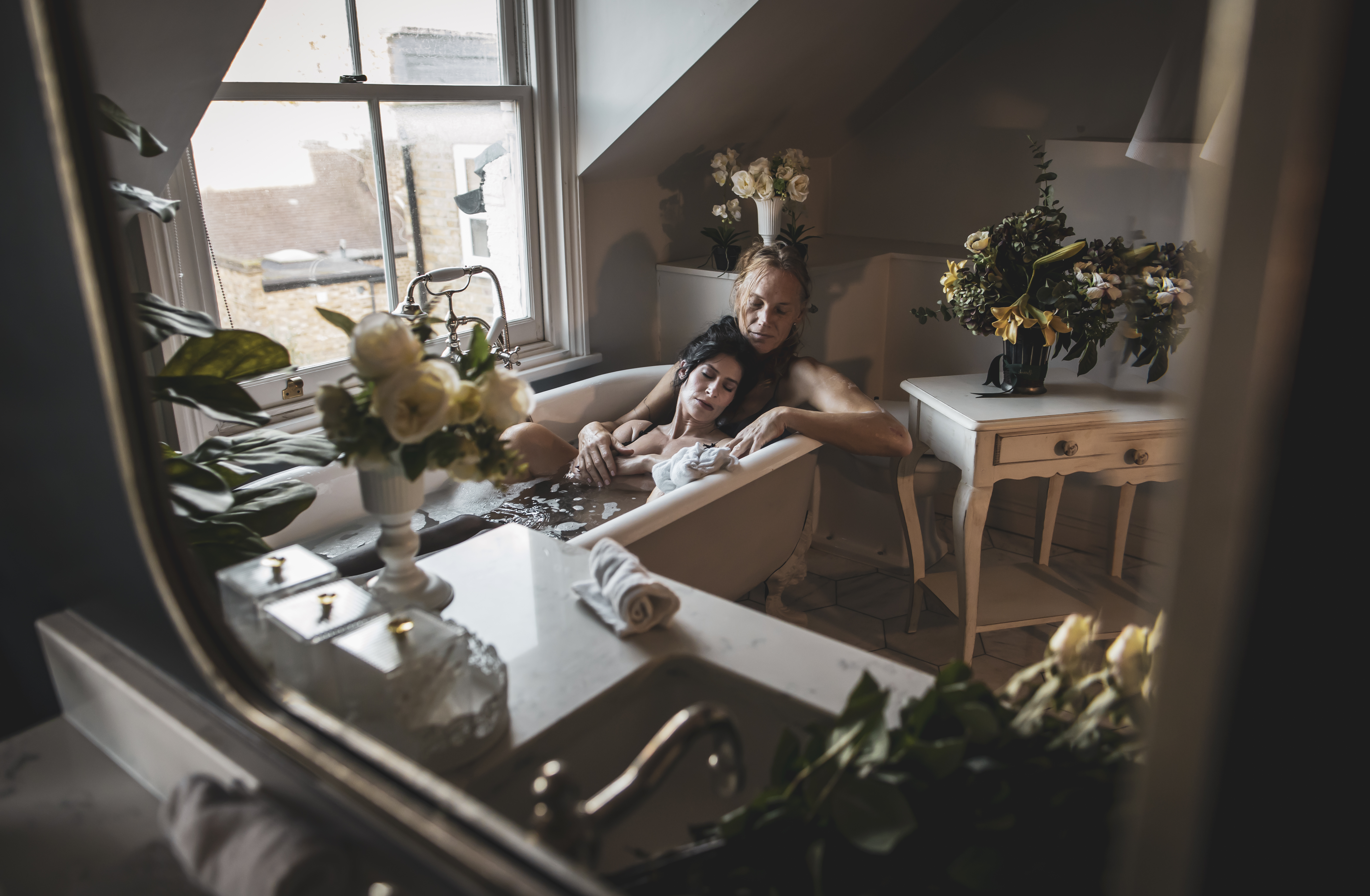 Valentina Dellagravis holding a woman in a warm bath in an intimate private setting with flowers and soft light.
