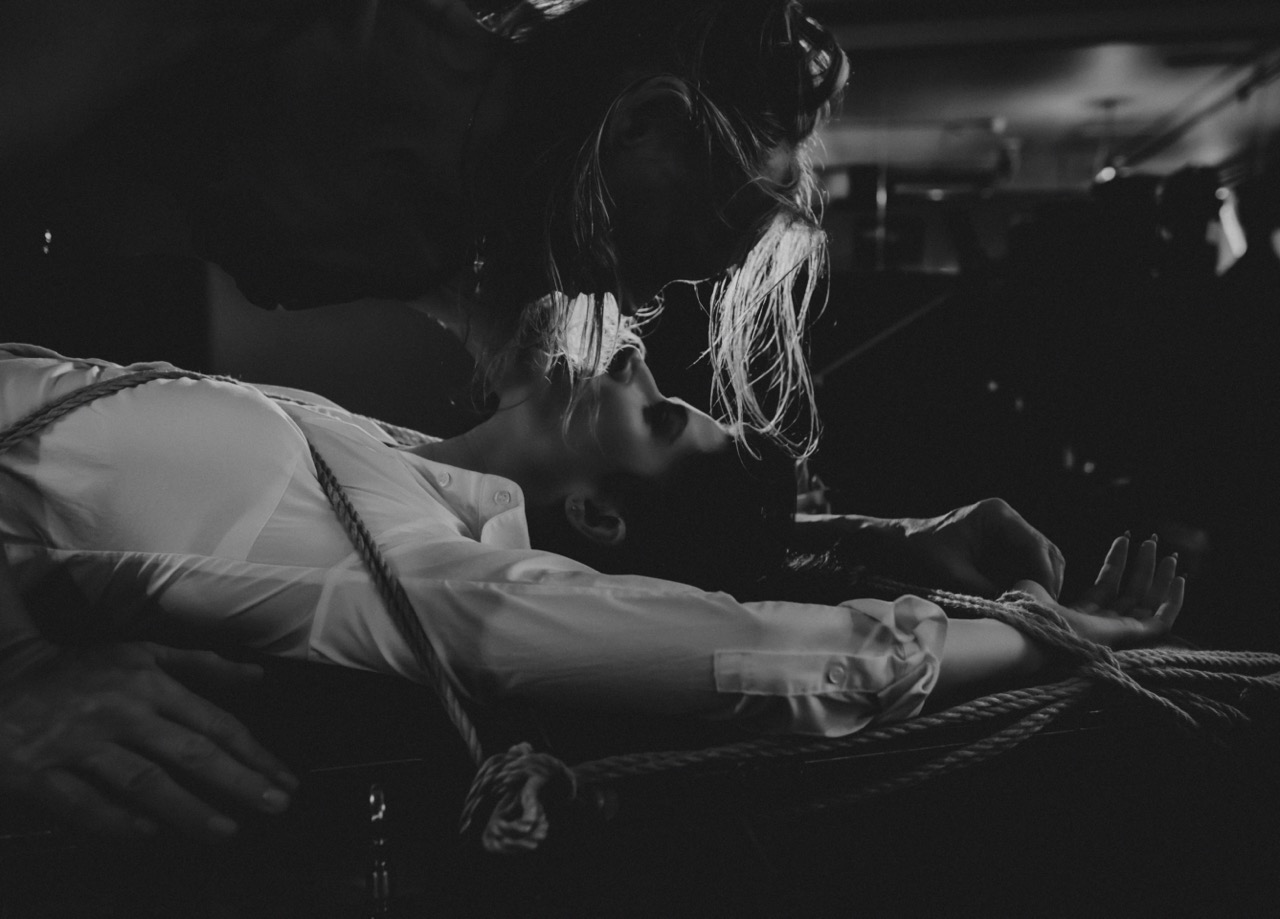 A bound submissive lies back as a dominatrix leans closely over them during a BDSM scene.